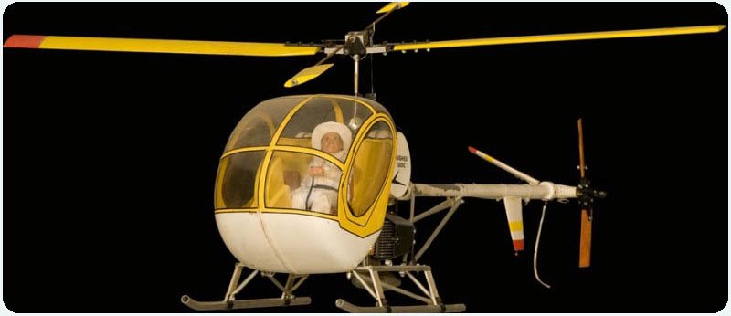 Close up shot of a yellow and white Hughes 3000 RC model helicopter. The canopy is clear and a pilot figurine in a white suite and hat can be clearly seen.jpg